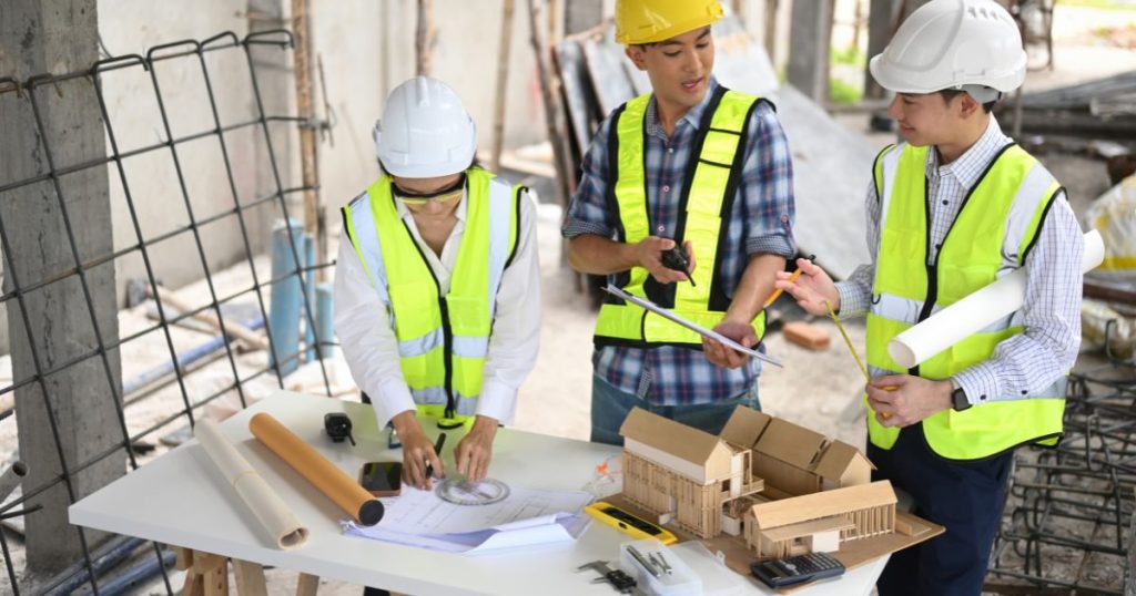 Drie personen die betrokken zijn bij een nieuwbouwproject: links een vrouw met witte veiligheidshelm, witte jas en fluohesje, in het midden een man met blauwgrijs geruit hemd, gele veiligheidshelm en fluohesje, rechts een man met gestreept wit hemd, witte veiligheidshelm, fluohesje en een opgerolde bouwtekening onder de arm. Op tafel liggen verschillende documenten en staat een maquette. 