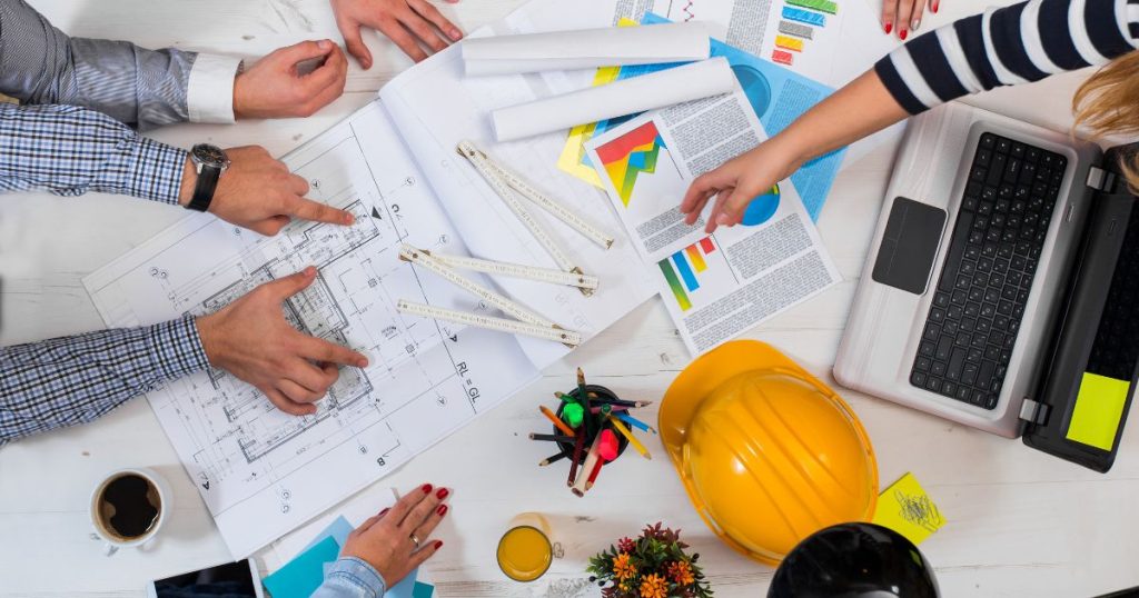 Een tafel met bouwplannen, papieren met grafieken, een laptop, een koker met schrijfgerei en een gele veiligheidshelm. Rondom de tafel de handen en armen van verschillende bouwprofessionals.