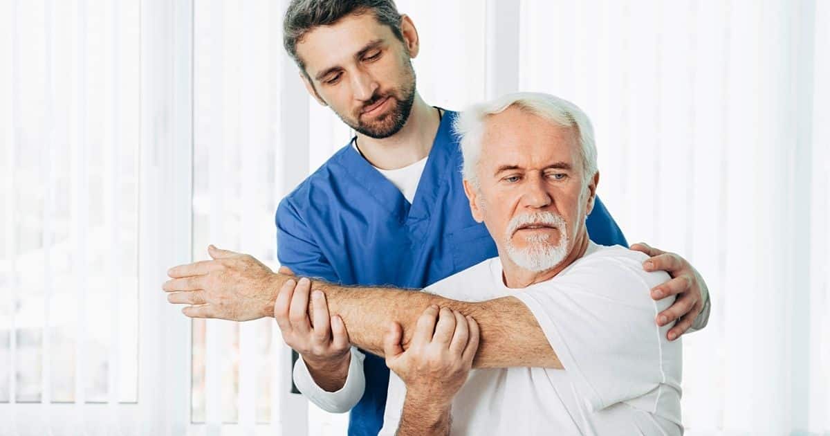 Een kinesist in blauwe tenue behandelt schouderklachten bij een man met wit haar en wit baardje in een witte t-shirt.