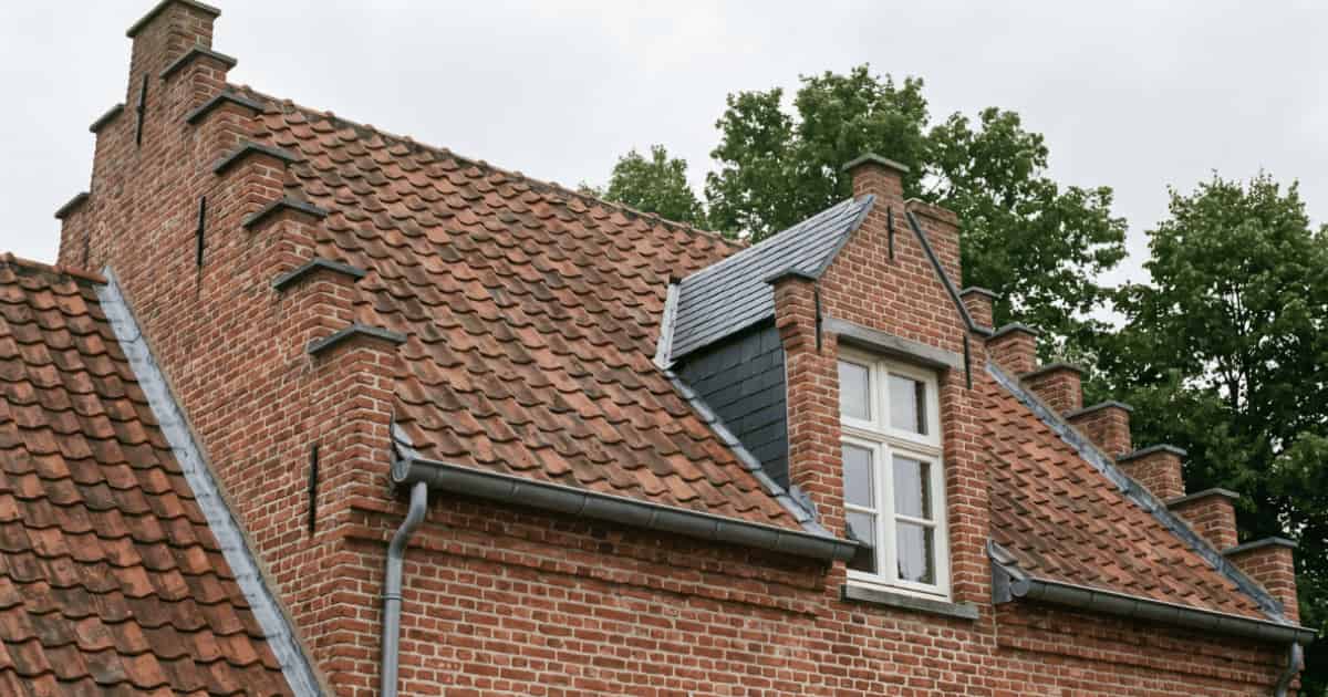 Een klassieke Vlaamse bakstenen woning met trapgevels draagt een hellend dak vol roodbruine Boomse pannen terwijl een dakkapel met donkergrijze leien bekleding en wit geschilderd houten raam uit het dakvlak steekt tegen een bewolkte lucht.