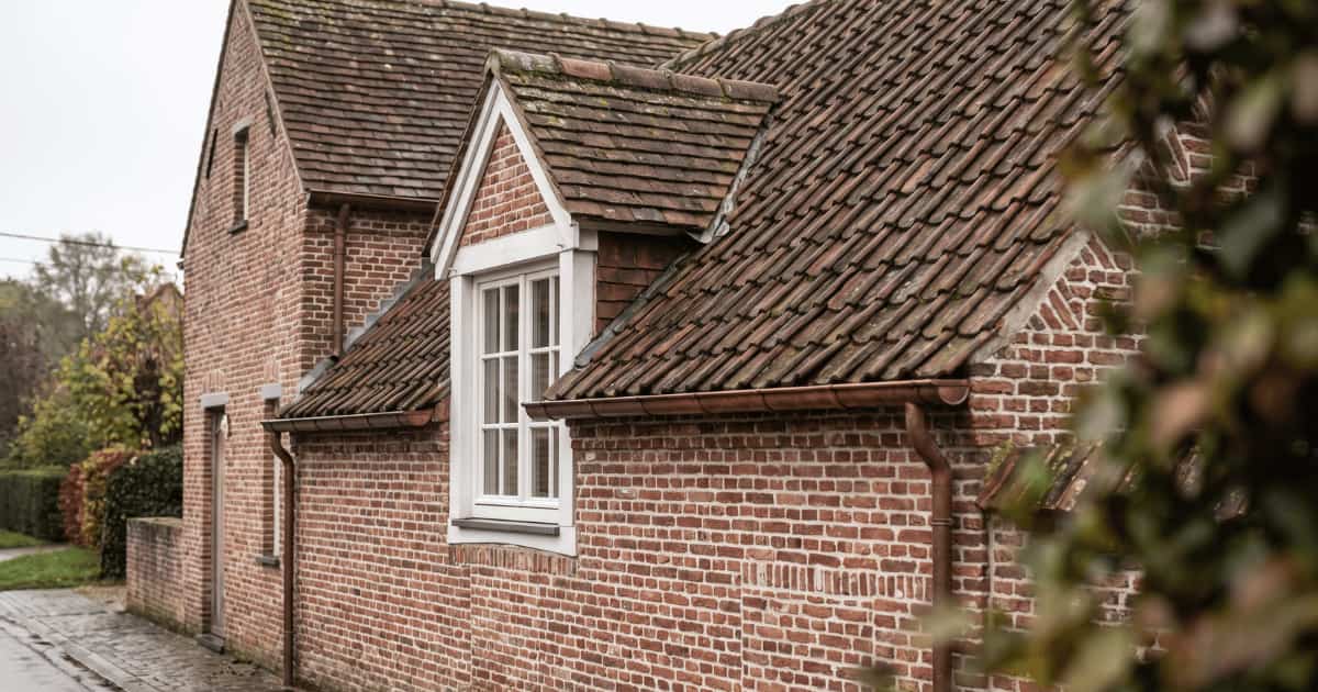 Een Vlaamse bakstenen woning met een puntdakkapel met witte houten raamkozijnen en een hellend dak bedekt met verweerde bruinrode kleipannen waar plaatselijk mos op groeit, met koperen regenpijpen langs de gevel onder een bewolkte hemel.