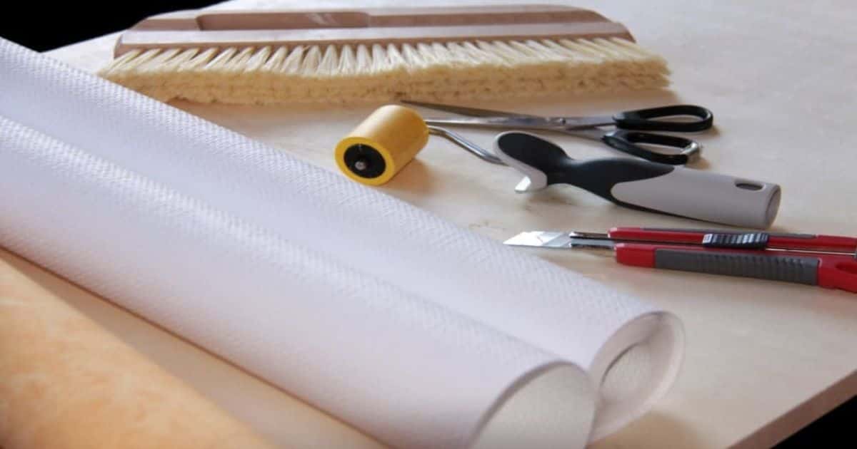 Vliesbehang op een houten tafel met werkgerief om het behang voor te bereiden en effectief te plaatsen: een breekmes, een schaar, een roller en een borstel.