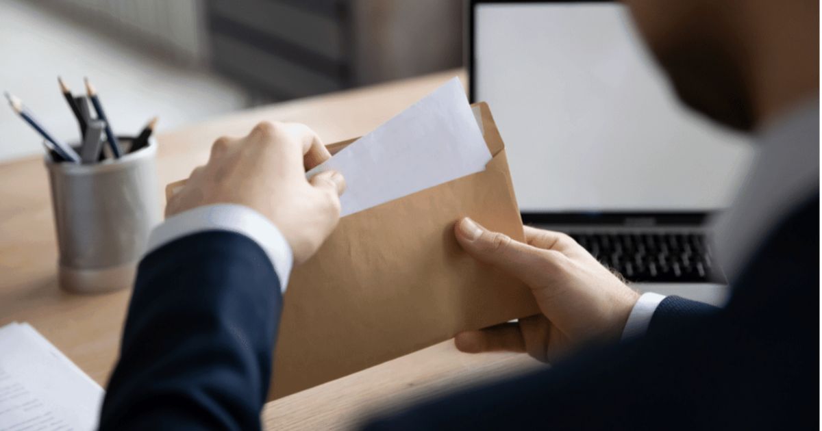 een man zit achter zijn bureau met zijn laptop opengeklapt en heeft een bruine envelop vast waar hij witte papieren uit haalt.