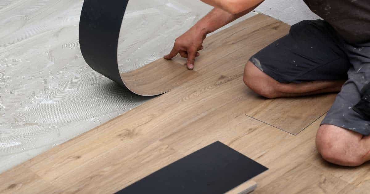 Een vloerspecialist kleeft smalle stroken vinyl met houtlook op een voorbereide ondervloer. Hij draagt een korte grijze werkbroek.