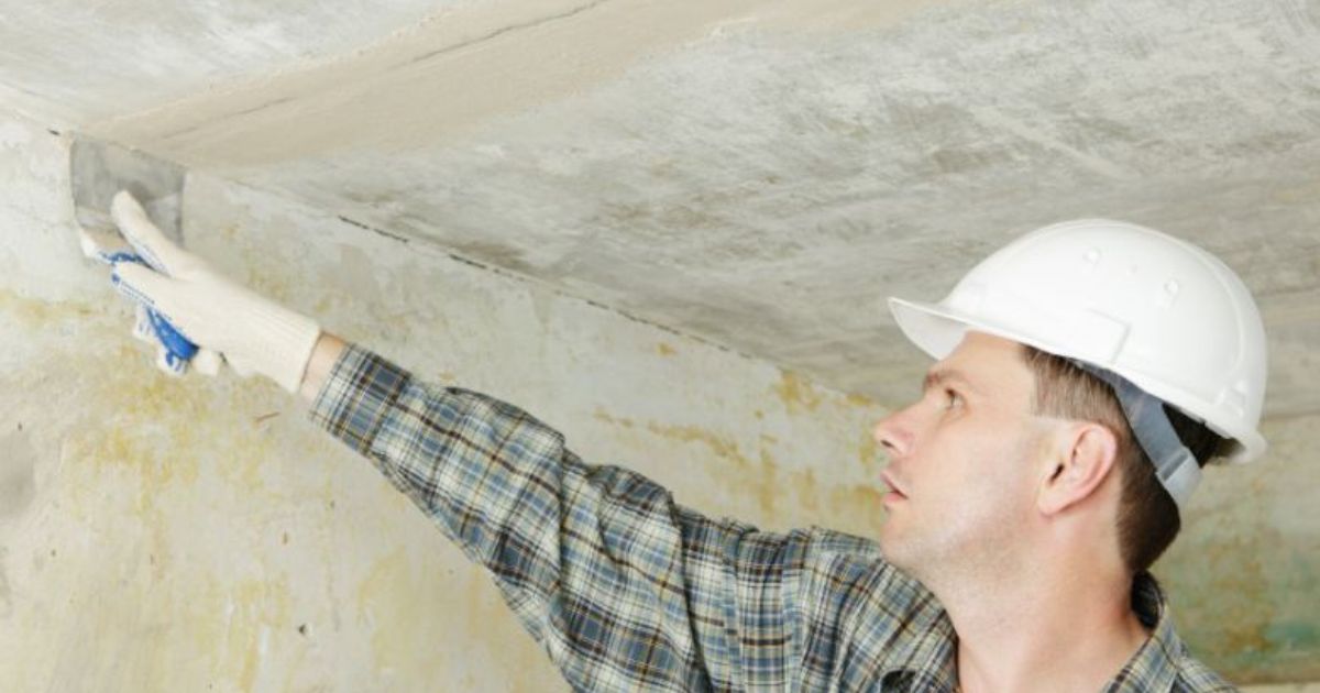 Een vochtbestrijder met ruitjeshemd en witte helm brengt met een spatel pasta aan op een muur die overduidelijk schade heeft opgelopen door vocht.