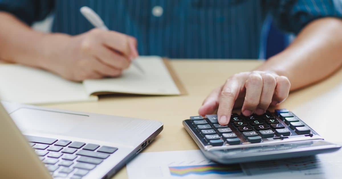 Een hand met de wijsvinger op een toets van een rekenmachine met afgeronde hoeken. De rekenmachine ligt op een document met grafieken, op een houten tafel. Naast de rekenmachine staat een laptop. Daarachter een notitieblok, met de andere hand, die ook een pen vast heeft.