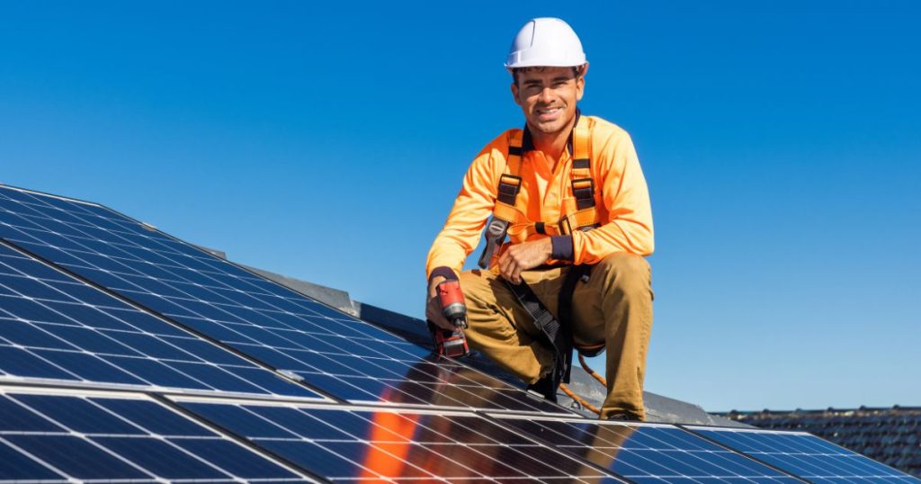 Een installateur met witte veiligheidshelm, oranje werkkledij en een boor in de hand zit op een dak met zonnepanelen.