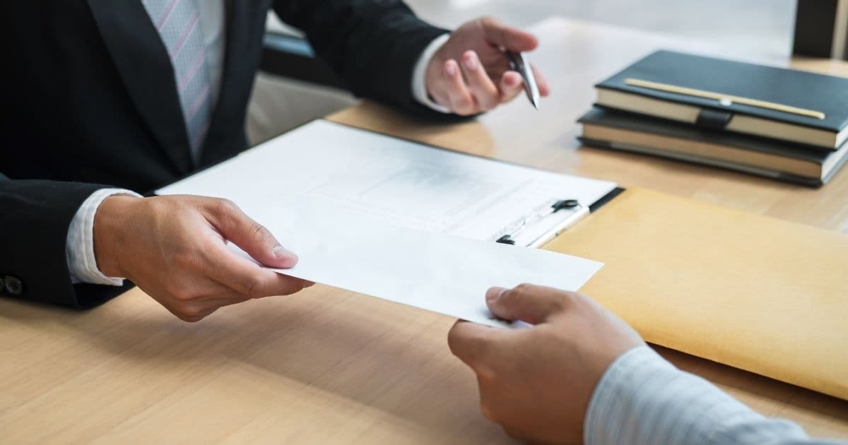 Een werknemer geeft zijn ontslag aan zijn werknemer middels een ontslagbrief. De brief zit in een witte enveloppe. Enkel hun handen en armen zijn in beeld: de werknemer draagt een blauw gestreept hemd, de werkgever een zwarte blazer, wit hemd en blauw met roze gestreepte das. Op de tafel tussen hen liggen een klembord, een bruine folder en twee zwarte notitieboekjes.