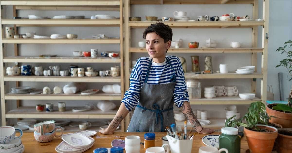 Een jonge dame met kort donkerbruin haar, een wit met blauw gestreepte trui en een grijs met blauwe schort staat in haar keramiekatelier. Achter haar staat een open houten kast vol keramische kopjes en borden. Voor haar, op een houten tafel, liggen nog meer van haar creaties. Er staan ook enkele kamerplanten in terracotta potten op de tafel.