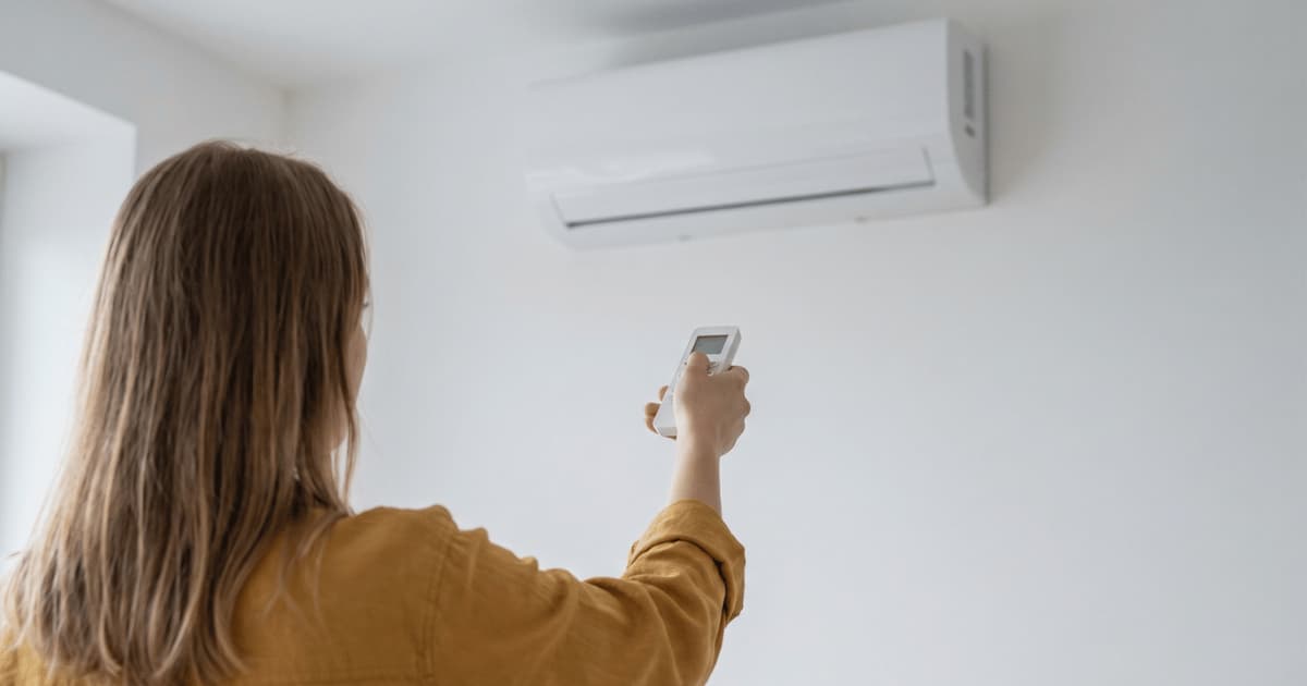 Een vrouw in een okerkleurig hemd richt vanuit de rug gezien een afstandsbediening op een witte airco-binnenunit die hoog op de witte muur hangt.