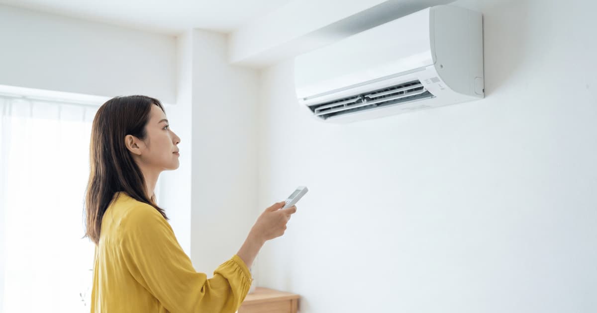 Een vrouw in een gele blouse staat in profielaanzicht en richt een witte afstandsbediening naar een wandgemonteerde airco-binnenunit met geopende lamellen in een lichte woonkamer.