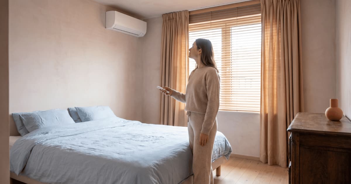 Een vrouw in beige loungewear staat naast een opgemaakt bed met lichtblauw beddengoed en richt een witte afstandsbediening naar de airco binnenunit die hoog aan de zandkleurige muur hangt, met warm licht dat door de houten lamellen van het raam valt.