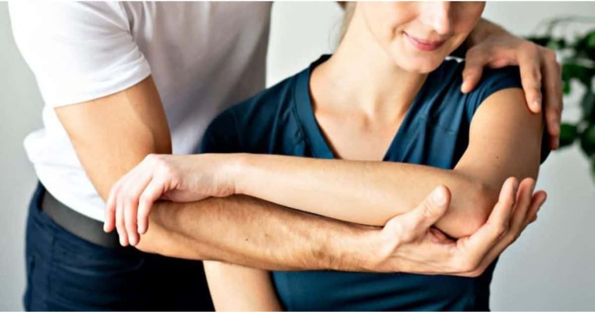 Osteopaat die aan het werk is bij een patiënte. Hij houdt haar linkerschouder vast met één hand en ondersteunt de linkerarm met een ander hand.