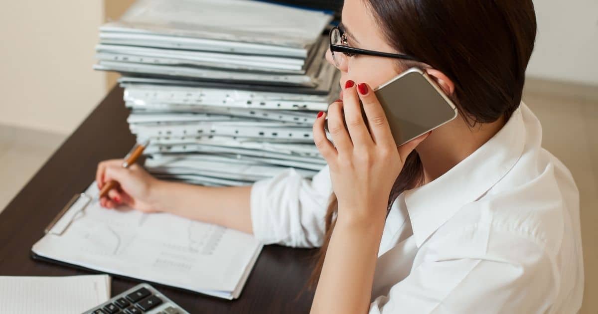 Een dame met een zwarte bril, bruin haar en wit hemd telefoneert terwijl ze met een pen iets noteert op een document met grafieken. Het document zit in een klembord. Links van het klembord ligt een rekenmachine, rechts een stapel dossiers in doorzichtige mapjes.