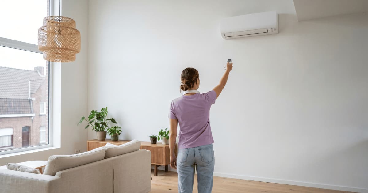 Een vrouw in een lila t-shirt en spijkerbroek staat met haar rug naar de camera en richt een witte afstandsbediening op een airco binnenunit hoog aan de witte muur in een lichte woonkamer met houten vloer, kamerplanten op een laag dressoir en bakstenen daken zichtbaar door het grote raam.
