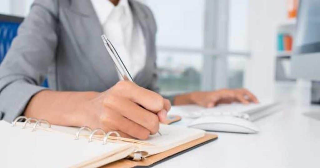 Een dame met grijs jasje schrijft met een balpen in een schriftje met ringen. Haar linkerhand houdt ze ondertussen op een wit toetsenbord.