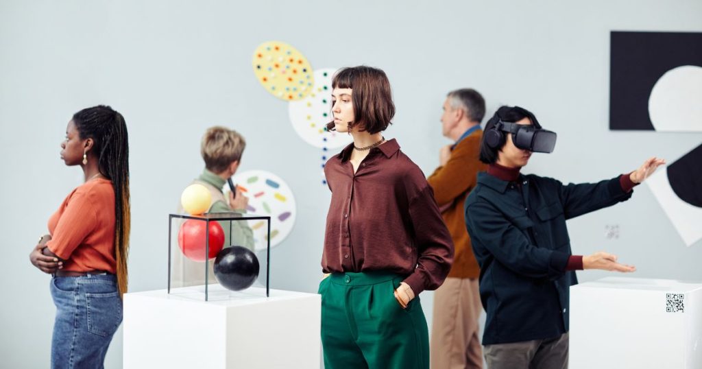 Een groep mensen in een museum met hedendaagse kunst. Een van de museumbezoekers draagt een VR-bril en bekijkt een virtueel kunstwerk met QR-corde.