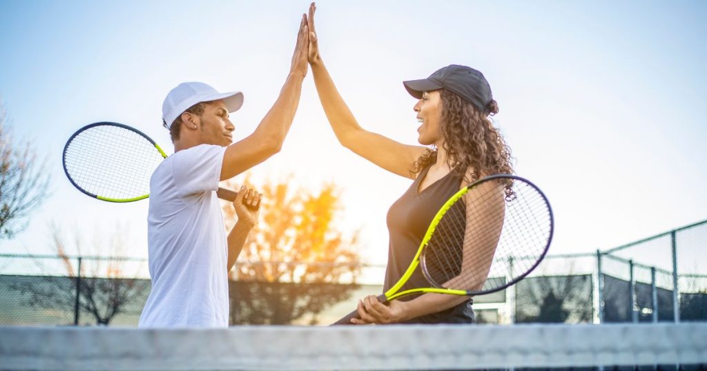 Twee mensen met tennisrackets en petjes geven elkaar een high five op een tennisveld.