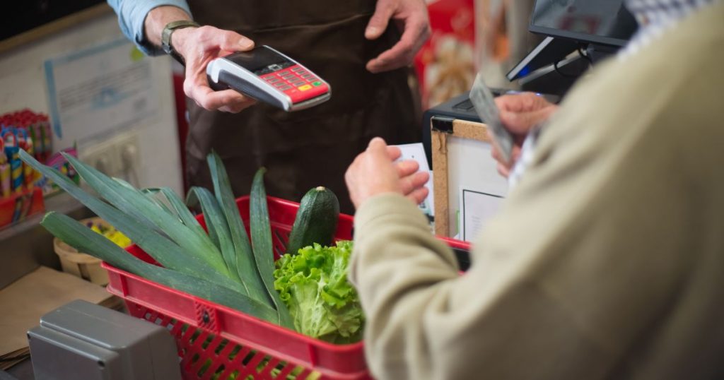 Een man in beige trui gebruikt een kaart om te betalen in de supermarkt. Voor hem staat een mandje met prei, een courgette en een krop sla. De winkelbediende, een man met bruin schort, blauw hemd en een horloge, biedt hem een betaalterminal aan.