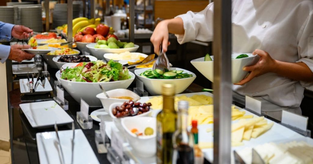 Een saladebar of buffet met witte kommetjes gevuld met groenten, fruit, sauzen, en flesjes olie en dressings. Een dame in wit keukenschort vult de kom met komkommers bij. Achteraan de bar schept een man in blauw hemd zijn bord vol. 
