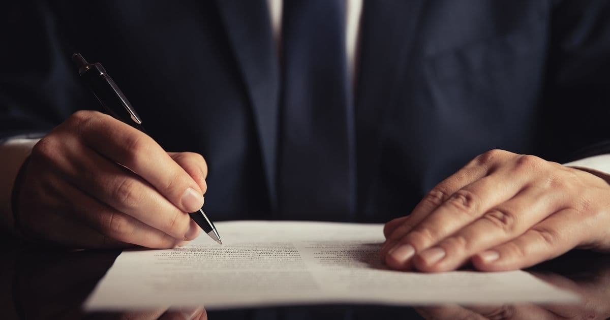 De borst en handen van een man met een wit hemd, blauwe das en blauwe blazer. In zijn rechterhand heeft hij een pen vast, de linkerhand ligt op een document met tekst.