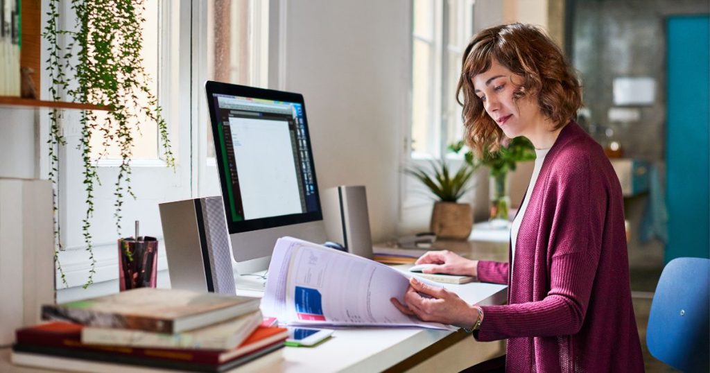 Een dame met paarse trui zit aan een tafel met computerscherm. Ze houdt een computermuis vast en bladert met een ander hand door een stapel papieren.