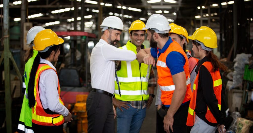 Een groep arbeiders in fluohesjes met veiligheidshelmen staat rond een man in zwarte broek, wit hemd en witte veiligheidshelm. De man in het witte hemd geeft zijn medewerker (een man in oranje fluohesje, blauw hemd en witte veiligheidshelm) een ellebooggroet. 