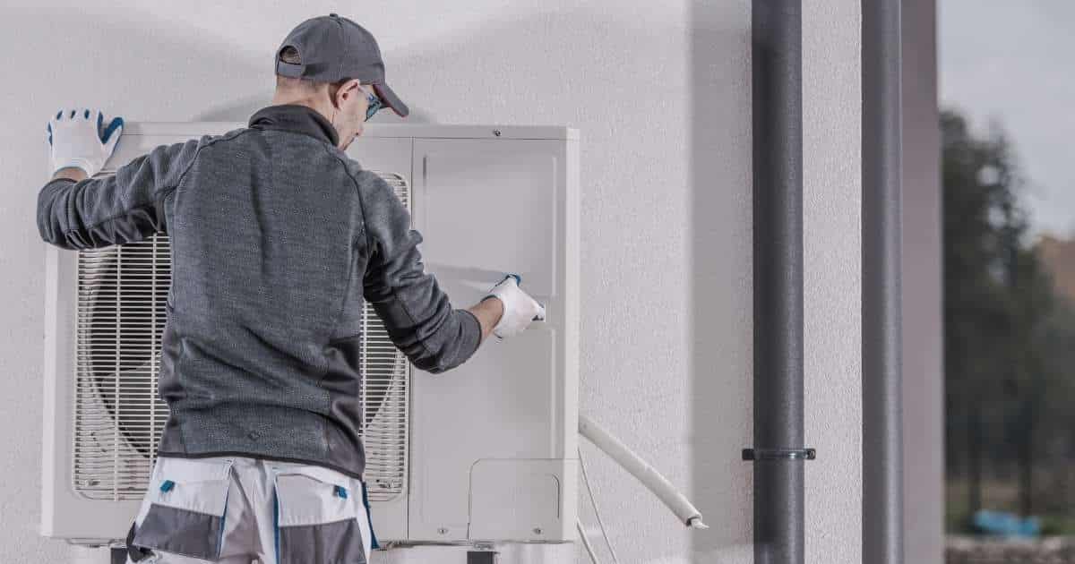 Een erkend installateur in grijze trui, grijze pet en lichtgrijze werkbroek plaatst een lucht-lucht warmtepomp tegen een witte gevel.