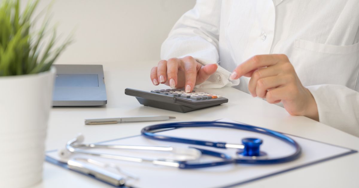 Een dokter gebruikt een rekentoestel. Naast hem of haar liggen een klembord met papier, een balpen en een stethoscoop. Ook staat er een laptop en een plant op de tafel.