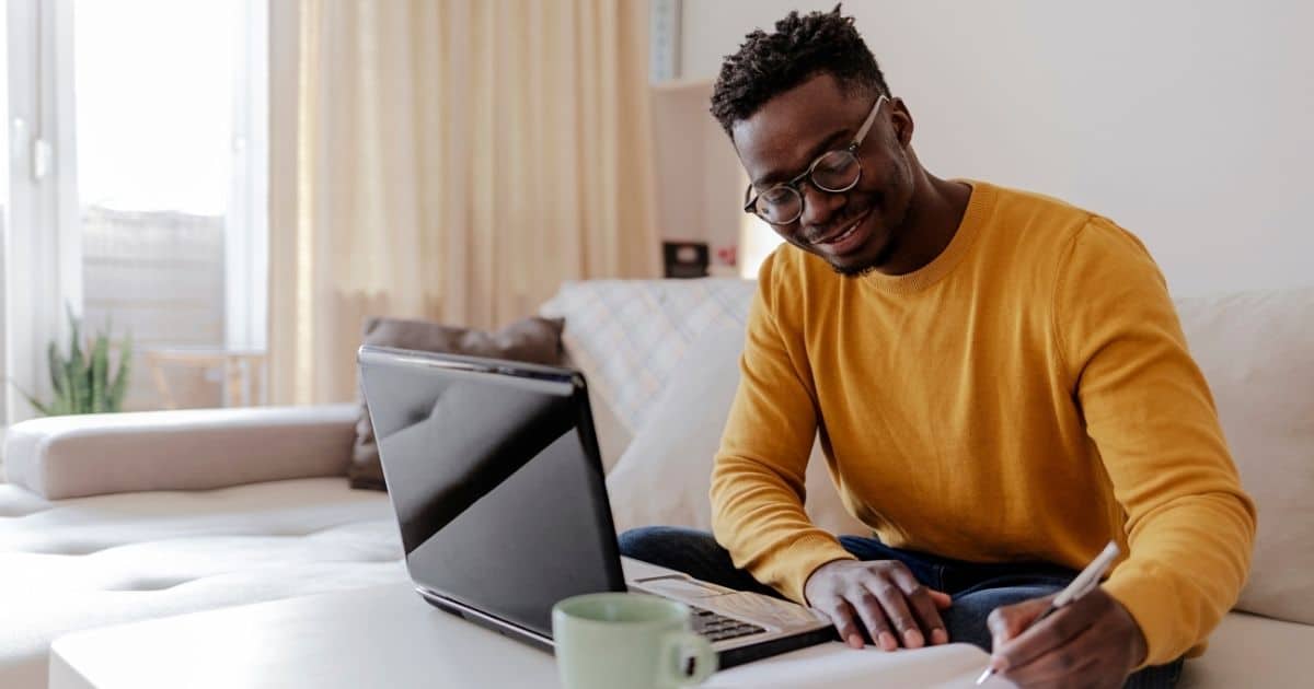 Een man met kort bruin haar, een bril met ronde glazen en een warmgele trui noteert iets in een notitieboekje. Voor hem staat een zwarte laptop op een witte tafel. Hij zit op een witte hoekzetel.