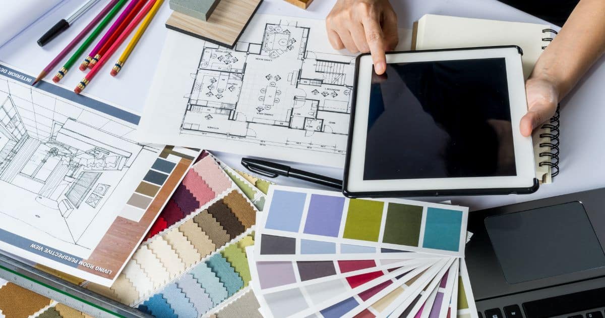 Het bureau van een interieurarchitect, bezaaid met verschillende waaiers met textuur- en kleurstalen, schetsen, ontwerpen, tekenmateriaal, notitieblokken en een tablet.