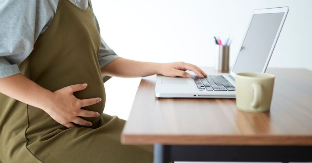 Een zwangere vrouw zit aan een laptop. Ze draagt een groene overall boven een grijs t-shirt.