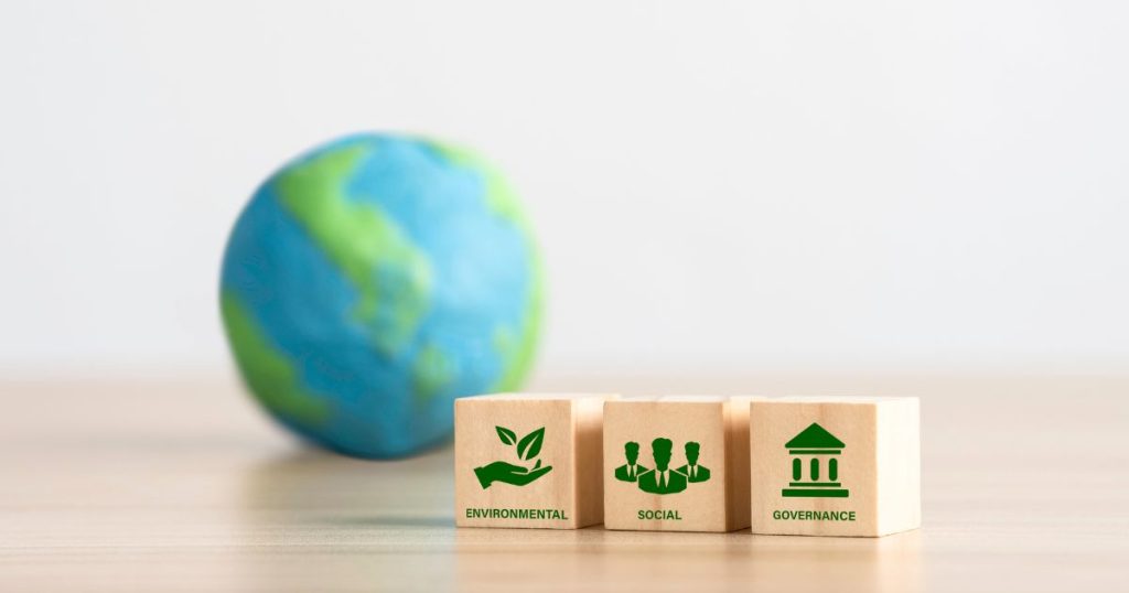 Een houten tafel met 3 houten blokken: 1 met een hand met bladeren boven en het woord 'environmental', 1 met drie mannenhoofden en -schouders en het woord 'social', 1 met een gebouw en het woord 'governance'. Op de achtergrond ligt een kleine, blauw met groene wereldbol.