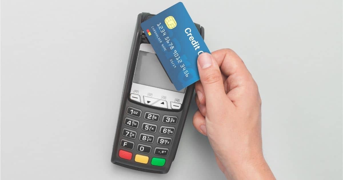 Een grijze betaalterminal tegen een lichtgrijze achtergrond. Een hand houdt een blauwe betaalkaart boven de terminal voor het uitvoeren van een contactloze betaling.
