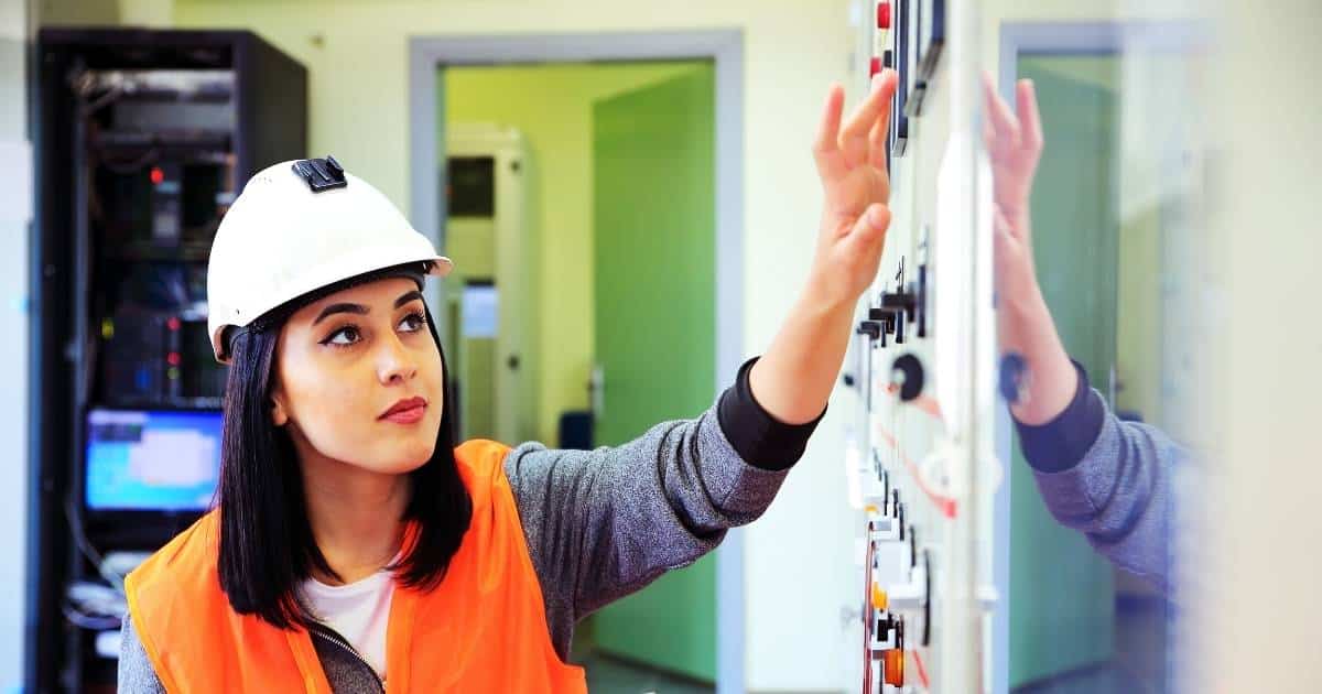 Een erkend keurder controleert een elektrische installatie. Ze draagt een grijze trui, oranje veiligheidsvest, en witte veiligheidshelm.