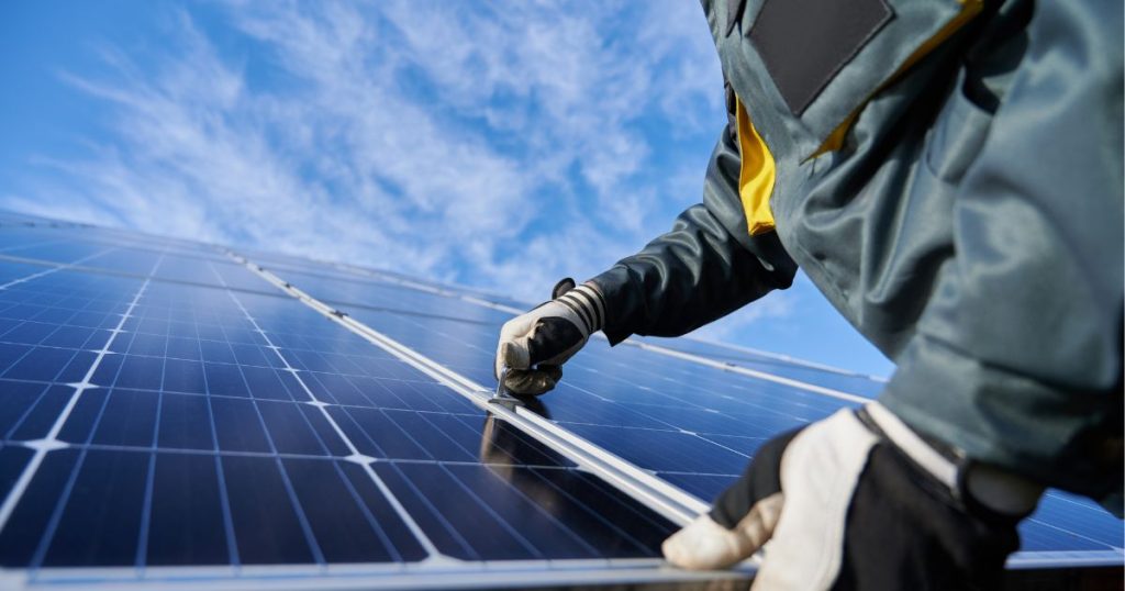 Iemand in een grijze jas en handschoenen monteert zonnepanelen op een dak