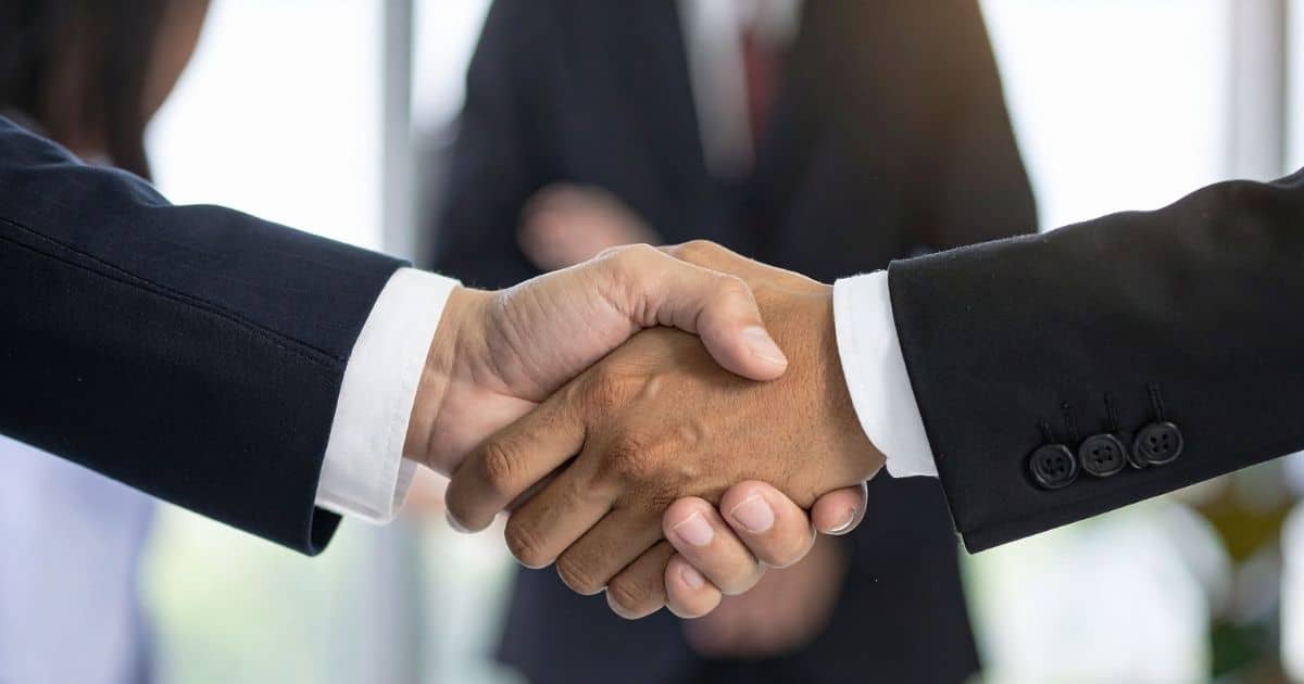 Twee mensen met witte hemden en zwarte vesten schudden elkaar de hand. Op de achtergrond, vervaagt, een derde figuur in maatpak.