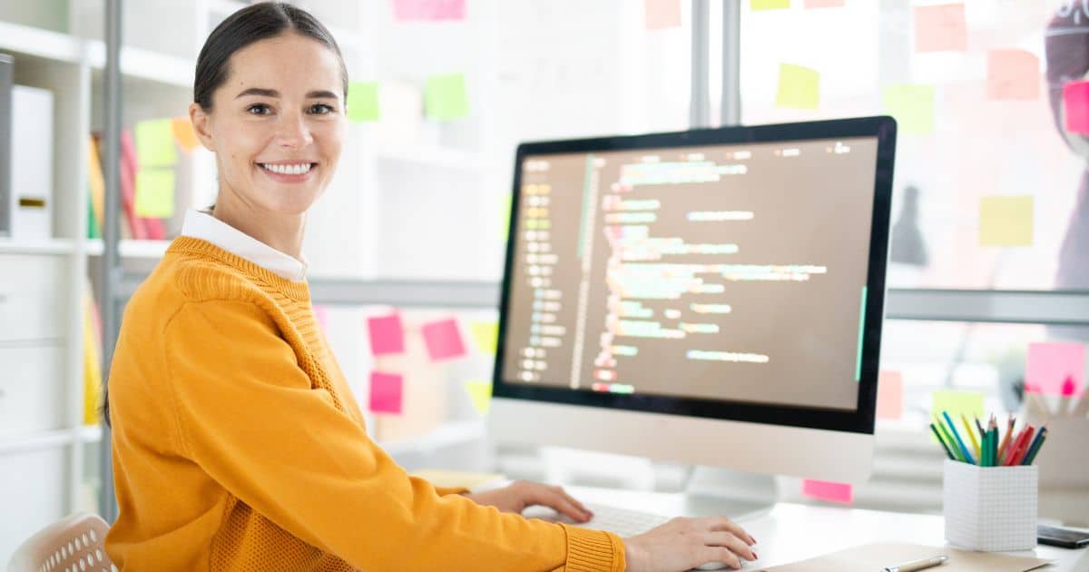 Een webdesigner zit voor haar computer. Op het scherm is ze bezig met het schrijven van de code voor een nieuwe site. Op het raam achter haar bureau kleven roze, oranje en gele post-its. Naast haar computer staat een wit bakje met schrijfgerei in allerlei kleuren.