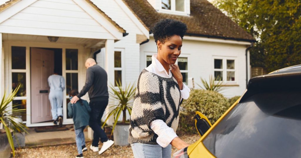 Een dame gebruikt een laadkabel van een bidirectionele laadpaal om haar gele elektrische auto op te laden. Op de achtergrond stappen een jonge vrouw, een man en een jongetje een woning met witte gevel binnen.