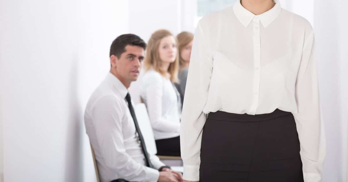 3 werknemers zitten op een stoel in een gang met witte muren. De achterste 2 zijn blonde vrouwen met elk een wit hemd, de man heeft kort bruin haar en draagt een wit hemd met zwarte das. Hij kijkt toe hoe een vierde collega, een vrouw met wit hemd en zwarte rok wiens hoofd niet zichtbaar is, naar de kijker toe wandelt.