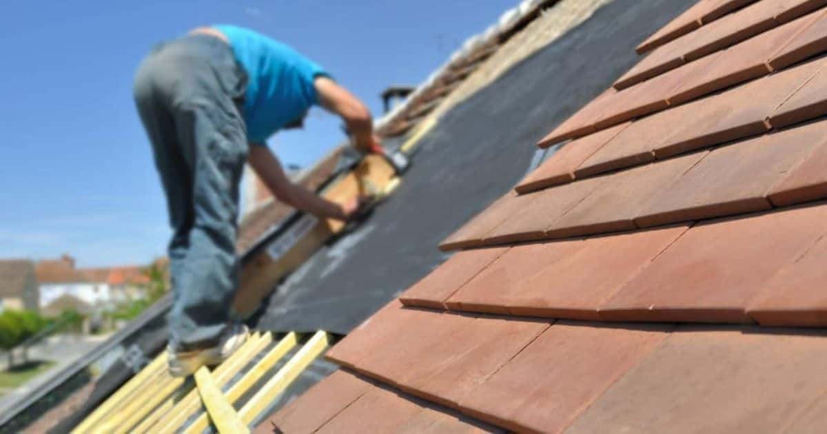 Een hellend dak met keramische tegelpannen. Een dakdekker in jeansbroek en blauw t-shirt steunt op de tengel- en pannenlatten terwijl hij het dakbeschot aanbrengt.