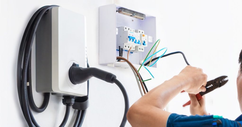 Een laadpaal naast een thuisbatterij. De arm van een installateur is in beeld: hij draagt een blauw werkhemd en heeft een tang vast.