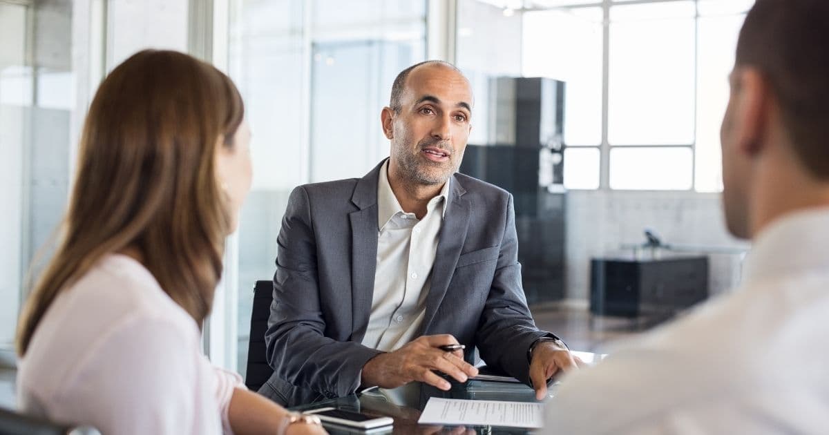 Een man in grijs pak en wit hemd zit aan tafel met een dame en een heer. Hij heeft een stoppelbaard en zeer kort haar. In zijn rectherhand heeft hij een pen vast. De dame, links voor de man, heeft bruin haar en draagt een lichtroze hemd. De heer, rechts voor de man, draagt een wit hemd en heeft kort haar en een lichte stoppelbaard. Op de houten tafel tussen hen liggen een smartphone en een blad.