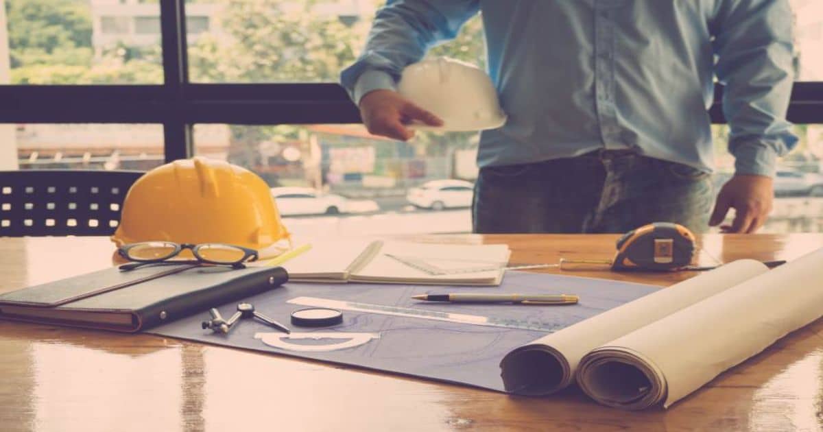 Werktafel met bouwplannen, veiligheidshelmen en meetgereedschap, met een persoon op de achtergrond.