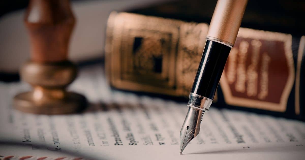 Een vulpen boven een officieel document. Op de achtergrond, vervaagt, een dik oud boek en een koperen stempel met houten handvat.