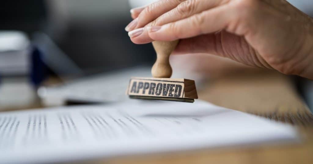 Een dameshand duwt een stempel met het woord 'approved' op een vergunningsaanvraag voor een uitbouw of aanbouw aan een bestaande woning.