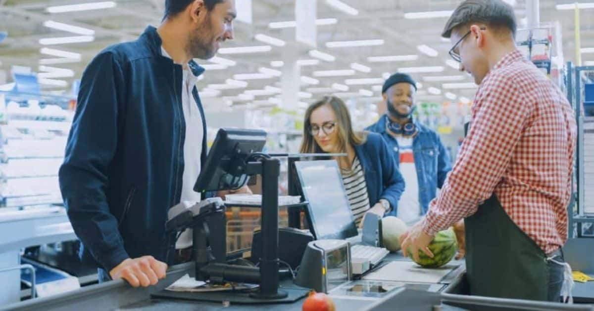 Een kassier en drie klanten staan rond het kassasysteem van een grote winkel. De kassier weegt een watermeloen met de ingebouwde weegschaal.