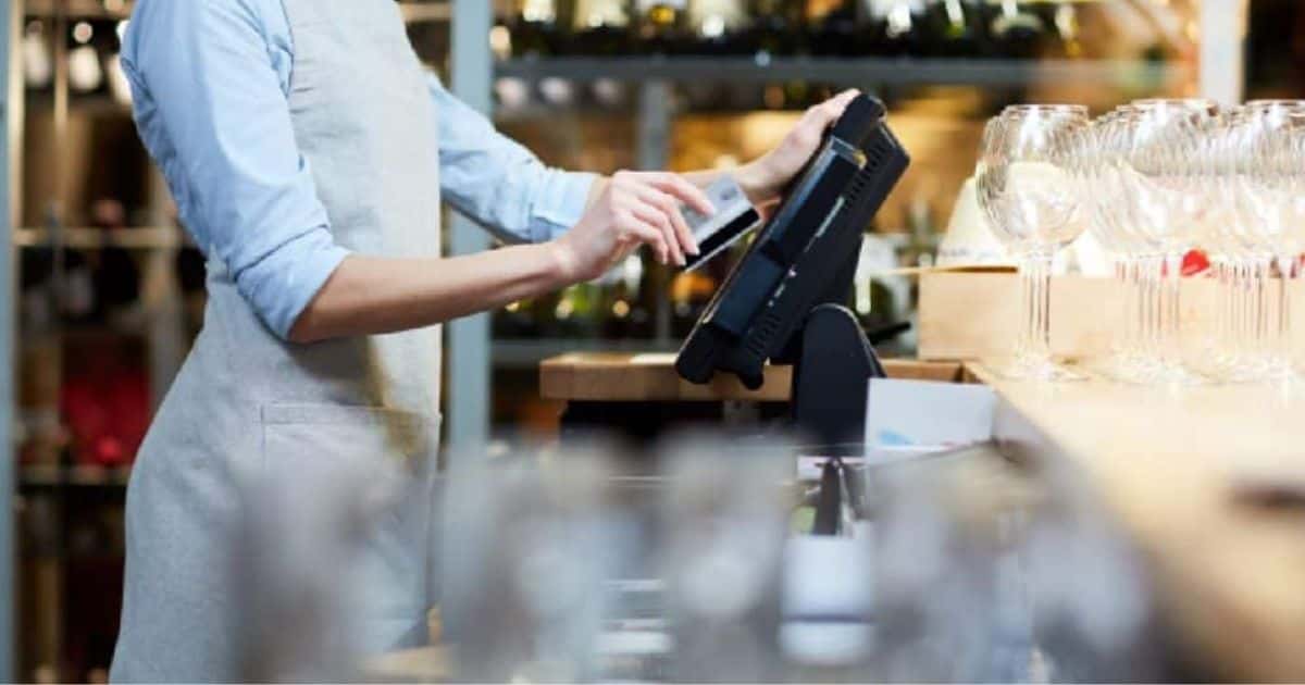 Een dame met een grijs schort gebruikt de betaalkaart van een klant om af te rekenen met het kassasysteem van haar horecazaak. Achter het kassasysteem staan enkele nette wijnglazen, en tegen de achterste muur staan rekken met wijnflessen.