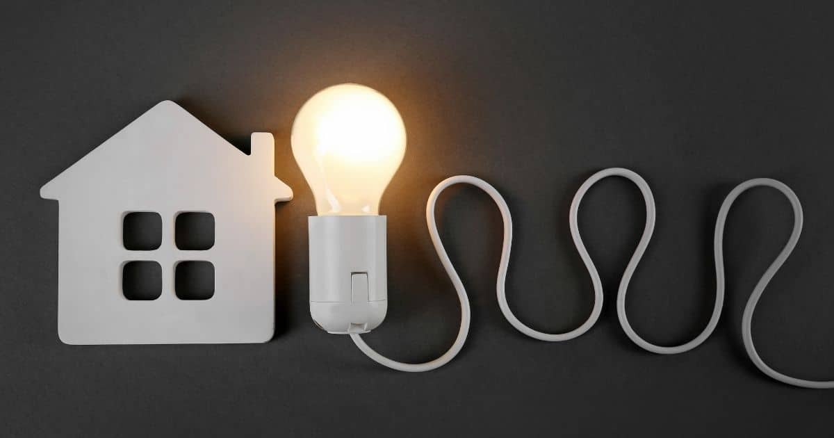 Een witte, simplistische voorstelling van een woning, met daarnaast een brandende witte lamp met snoer, tegen een matzwarte achtergrond.