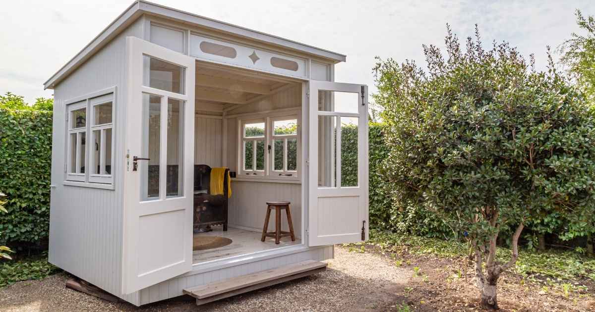 Wit houten tuinhuis met open deuren en een stoel binnen, naast een grote struik in de tuin.